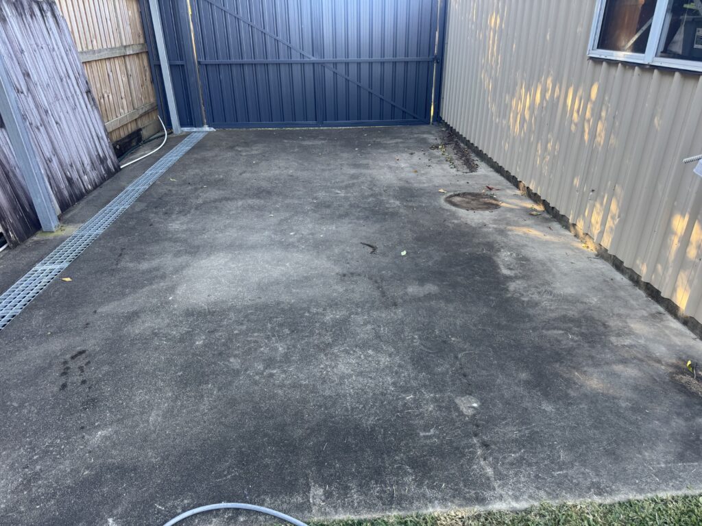 Concrete area beside a shed before cleaning, with dark surface build-up.